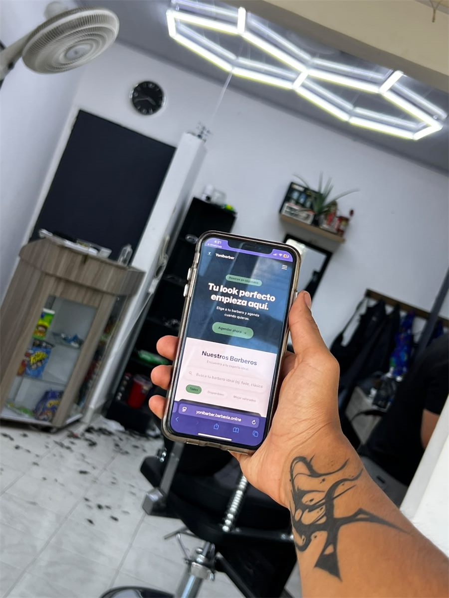 Celular dentro de la barbería Yonibarber mostrando su página de reservas con el mensaje "Tu look perfecto empieza aquí", con el local al fondo